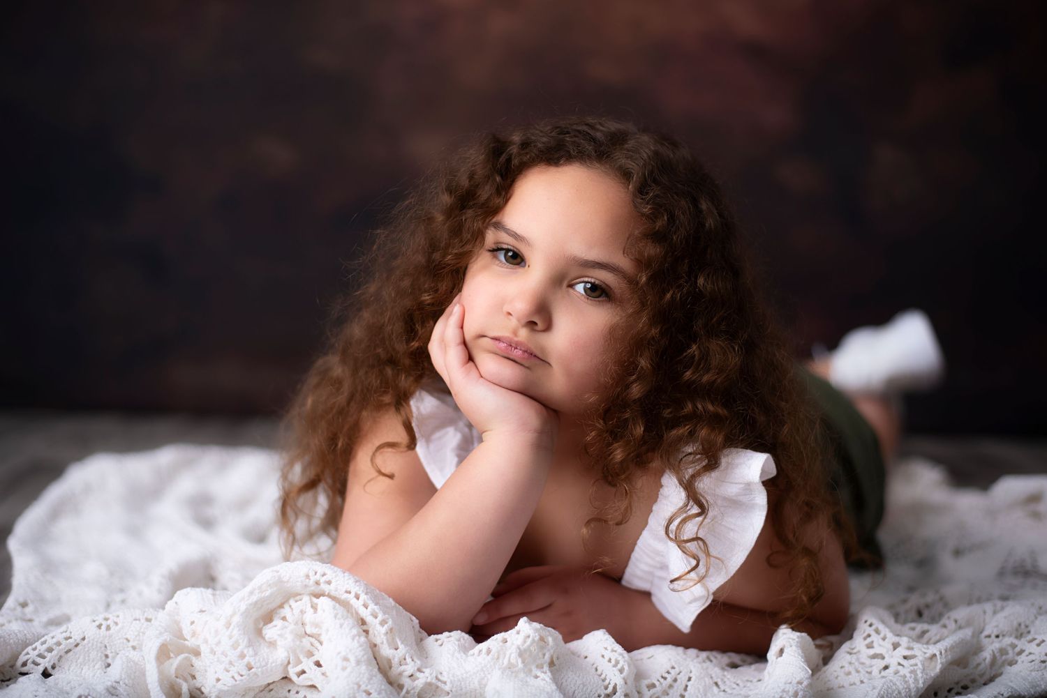 A young subject in a white lace dress lies on their stomach against a dark backdrop with curly hair cascading down.