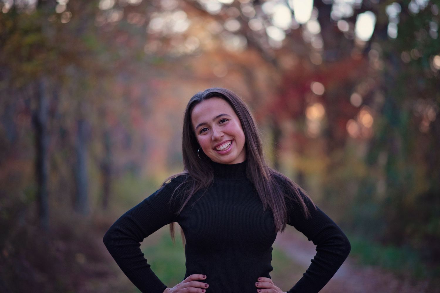 A person in a black turtleneck smiles outdoors against a blurred autumn backdrop with warm bokeh lighting.