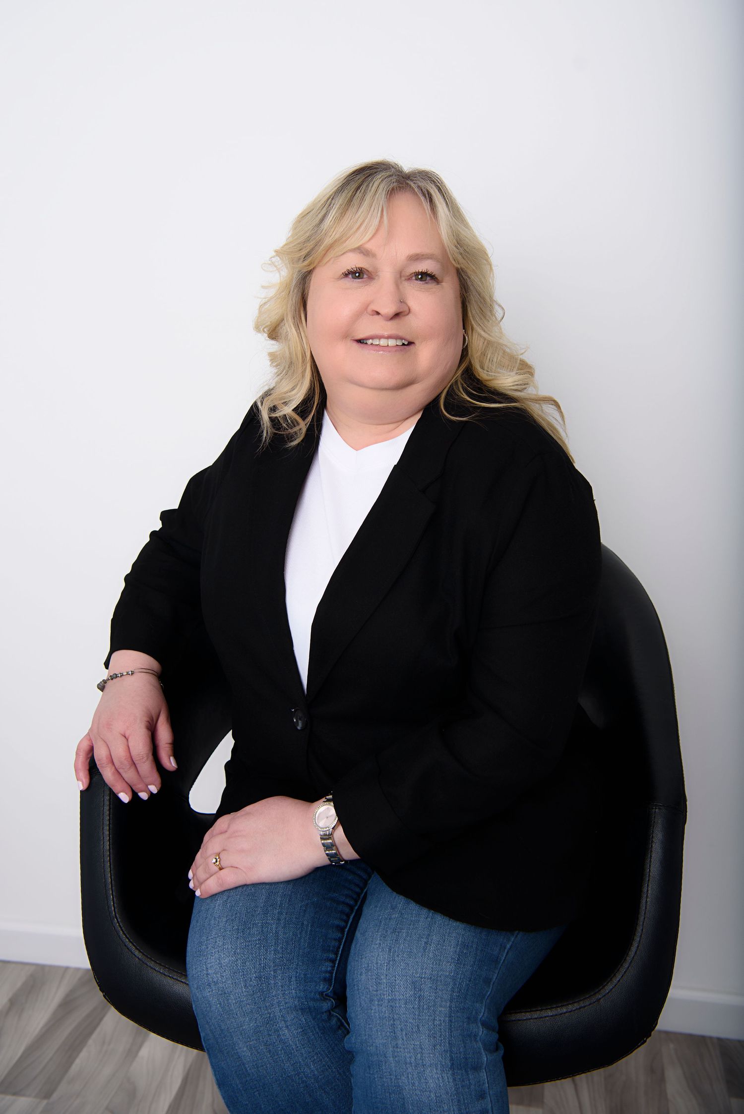 Professional portrait of someone in a black blazer and white top seated in a modern black chair against a light background.