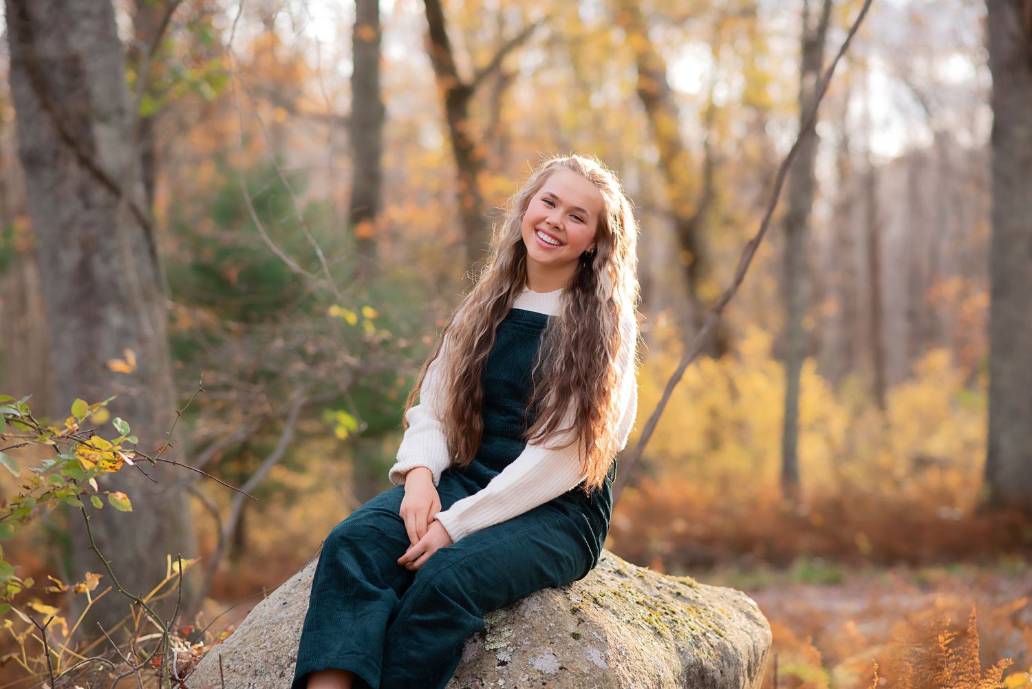 A person in a dark dress sits on a large boulder surrounded by autumn foliage in a wooded setting. Rhode Island Photographer