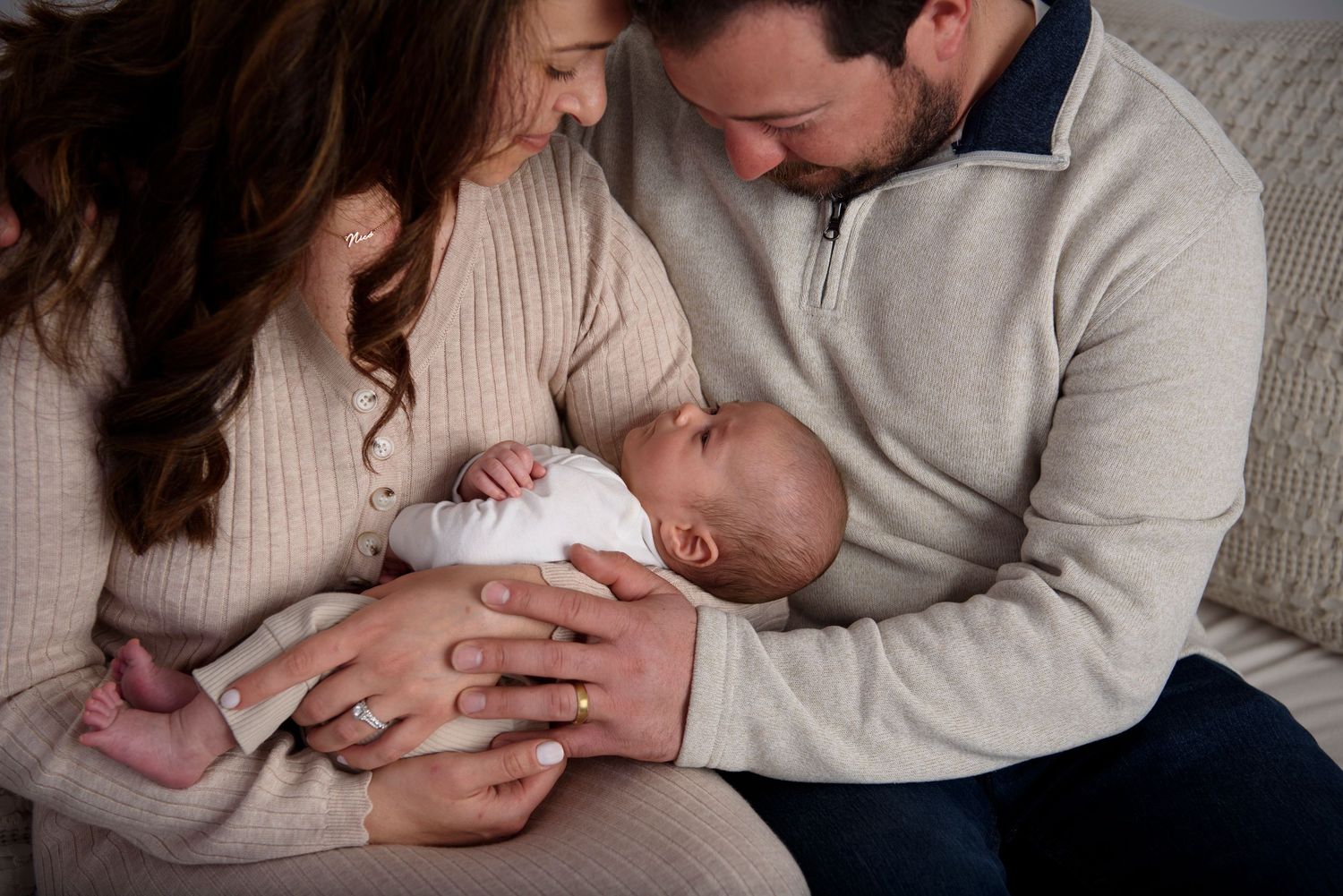 Two parents in cozy sweaters gaze lovingly at their sleeping newborn baby on a beige sofa.