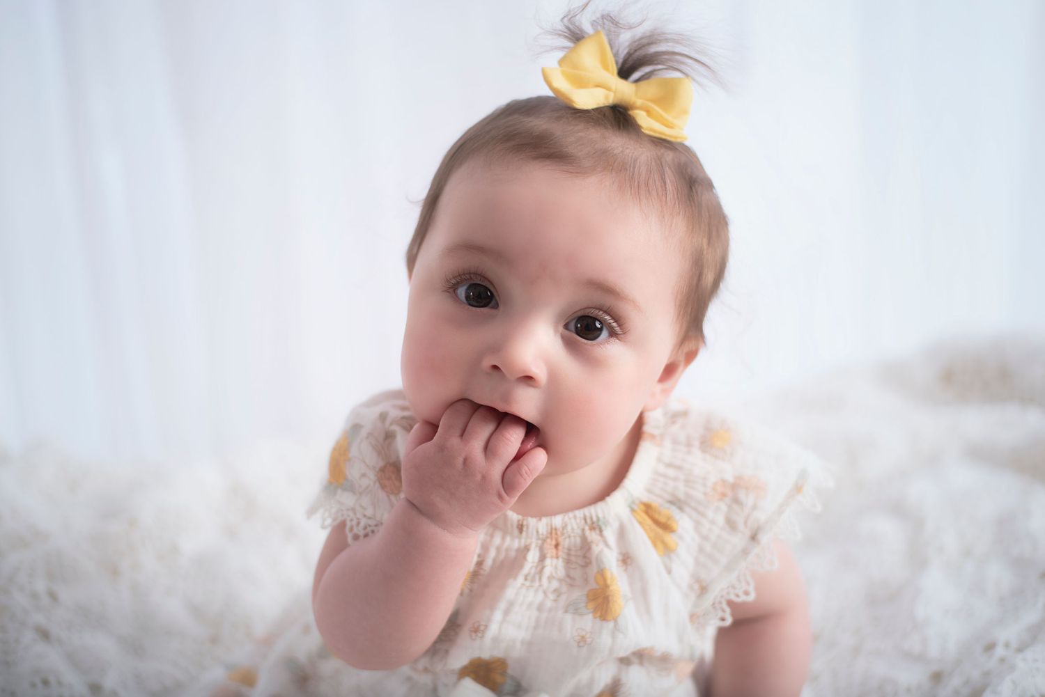Baby wearing yellow bow and white lace dress lying on white blanket while making playful expression.