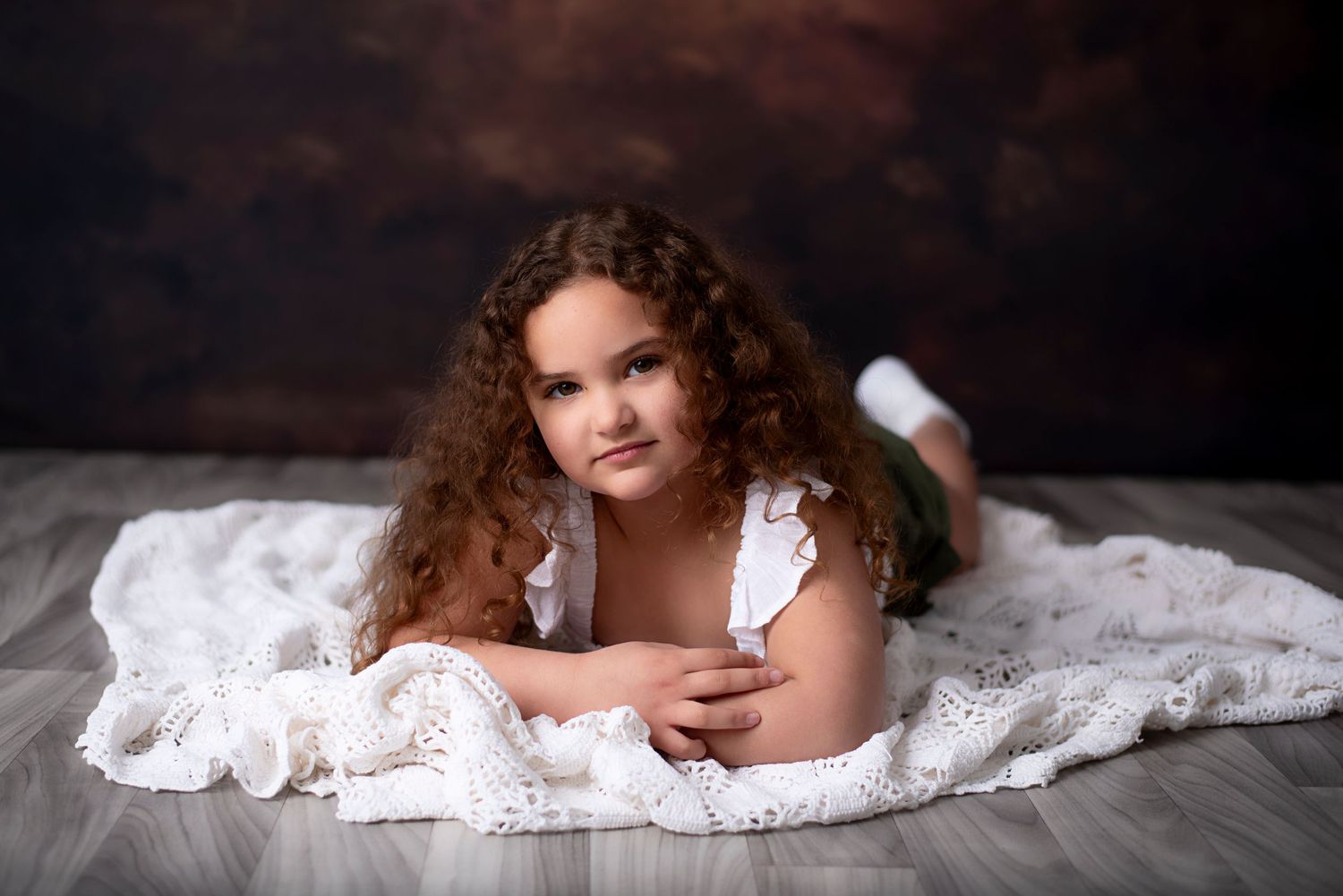 An infant in a soft white blanket lies down while looking at the camera in a studio setting.