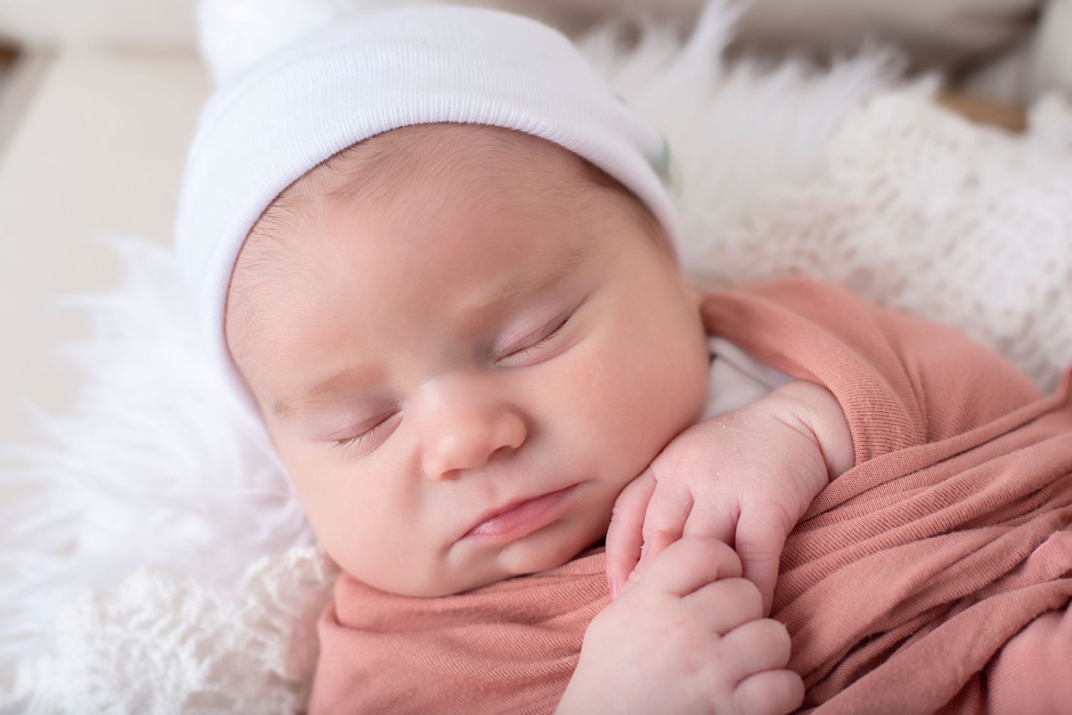 Peaceful sleeping newborn baby wrapped in soft pink fabric against white textured backdrop.