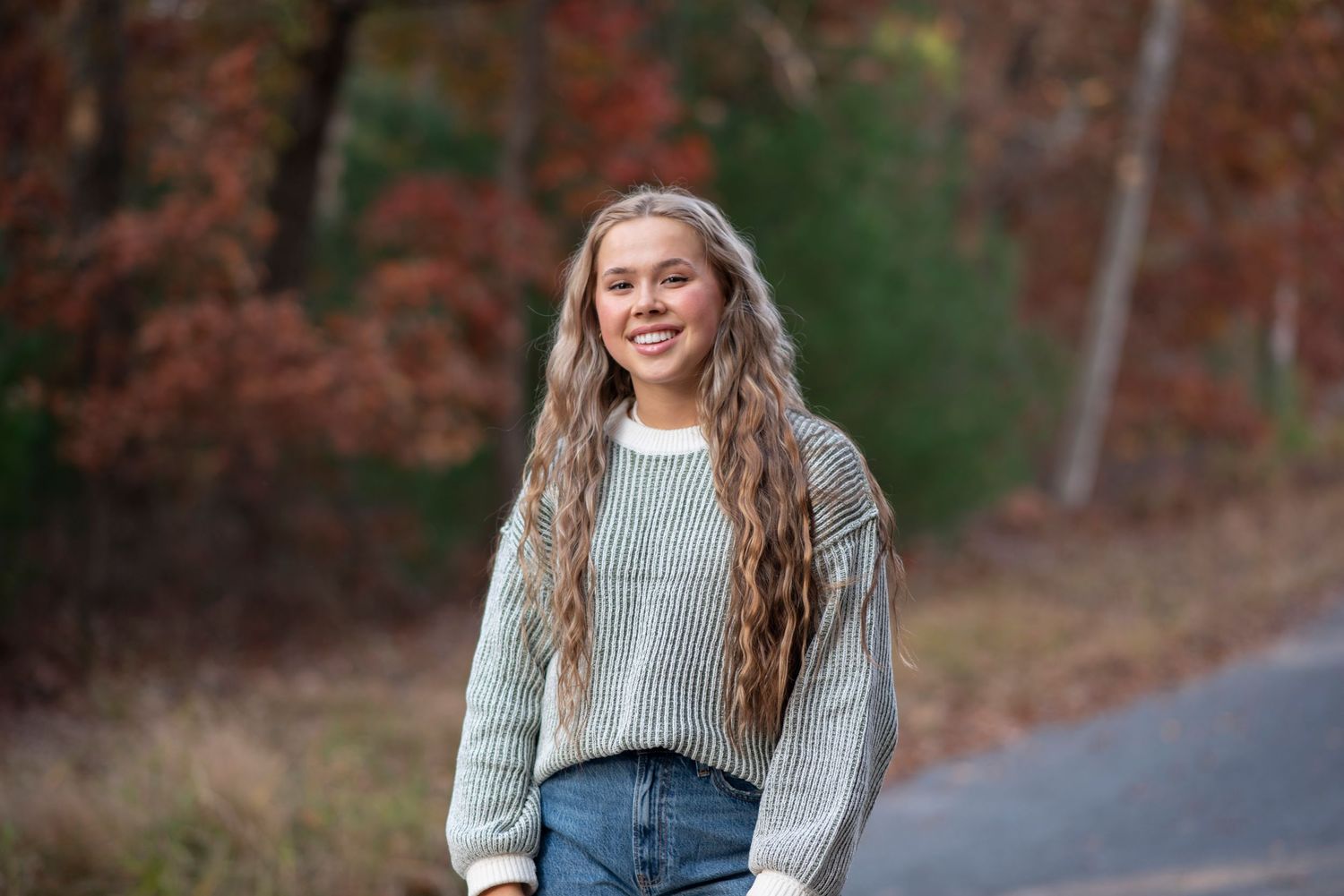 Person wearing a gray knit sweater and jeans smiles while standing on a road with autumn trees in background. Rhode Island Photographer