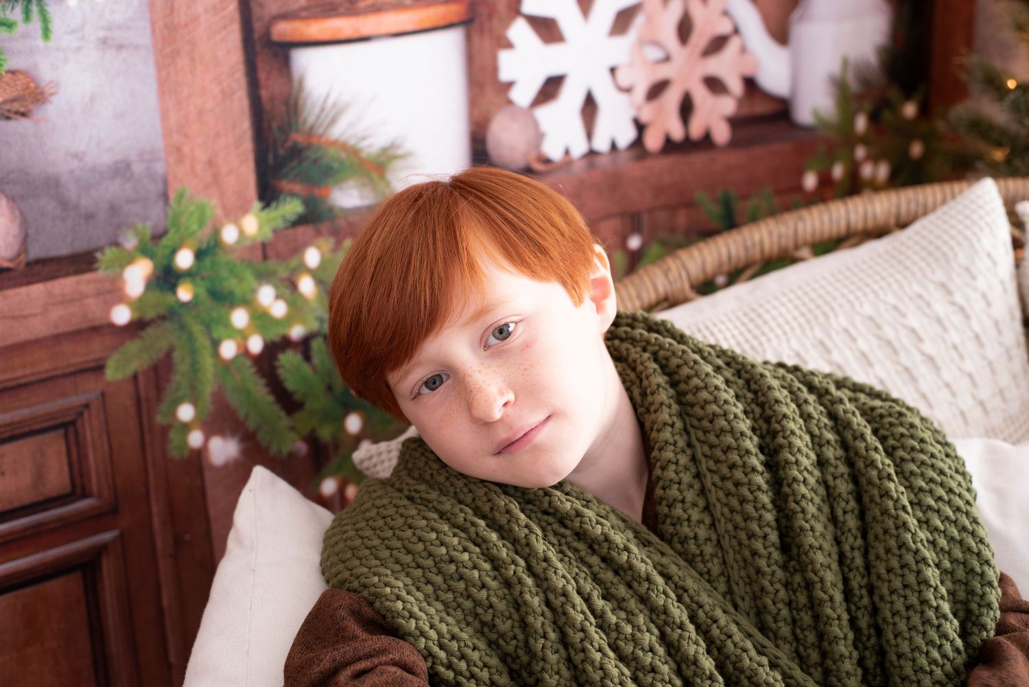 A person with bright red hair wearing a cozy olive green knit blanket sits against white pillows near winter decorations.