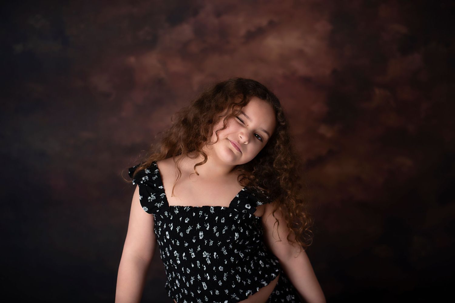 A person with long wavy hair against a dark stormy background wears a black floral dress.