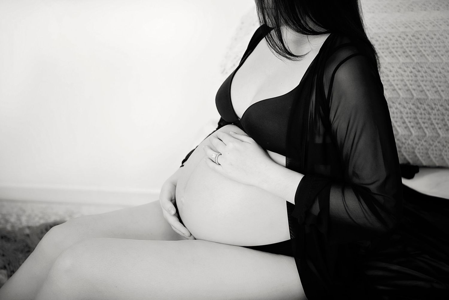 Black and white maternity photo shows expectant mother wearing sheer black top while sitting on bed.