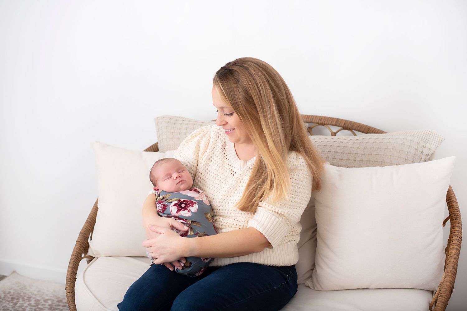 Woman in cream sweater sitting on rattan couch holds baby wearing striped outfit while surrounded by white pillows. RI Newborn Photographer