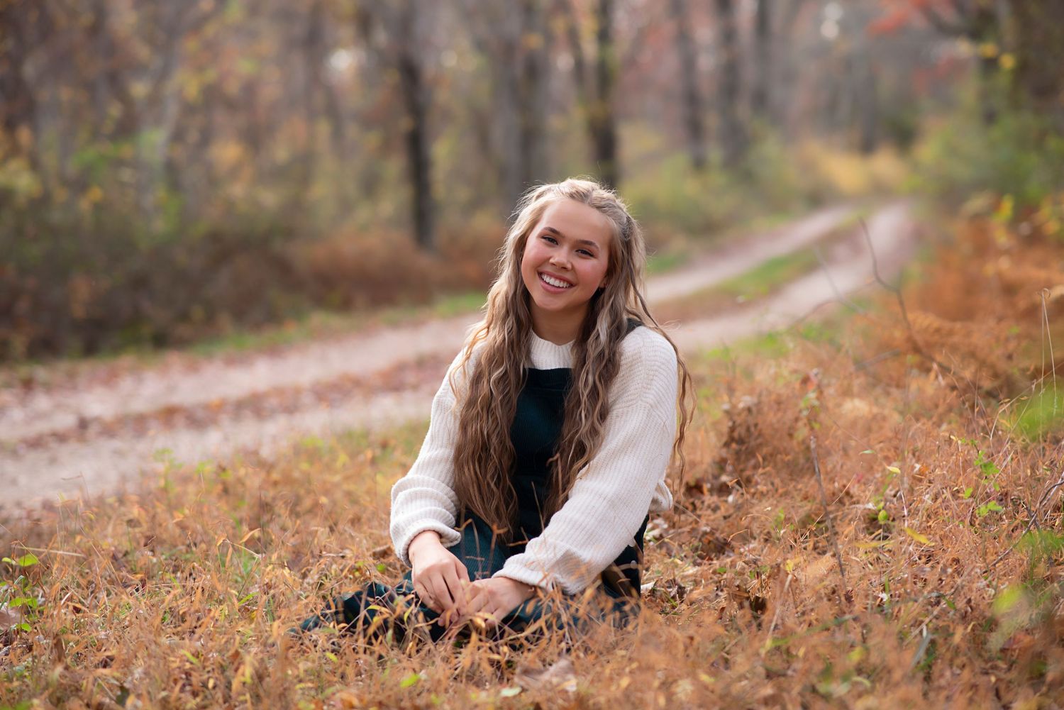 A figure seated in autumn foliage along a winding forest path wearing a cozy cream sweater. Rhode Island Photographer