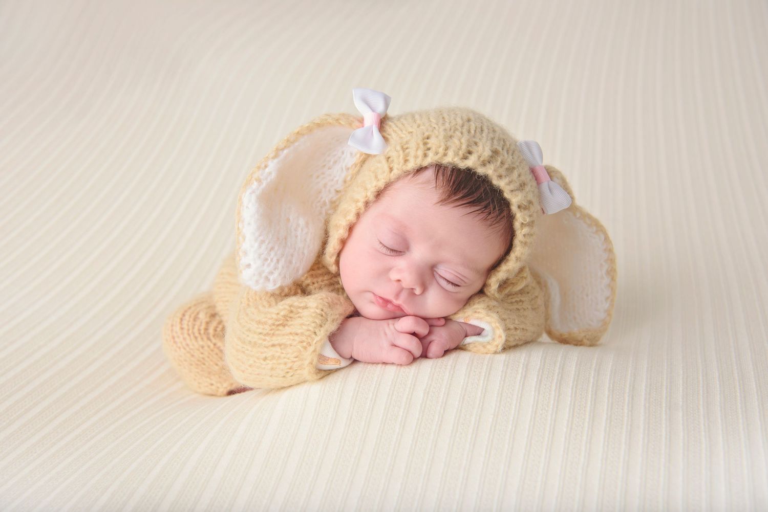 A sleeping newborn wearing a cozy knitted bunny outfit with ears lies on a cream-colored blanket. Exeter, RI Newborn Photographer