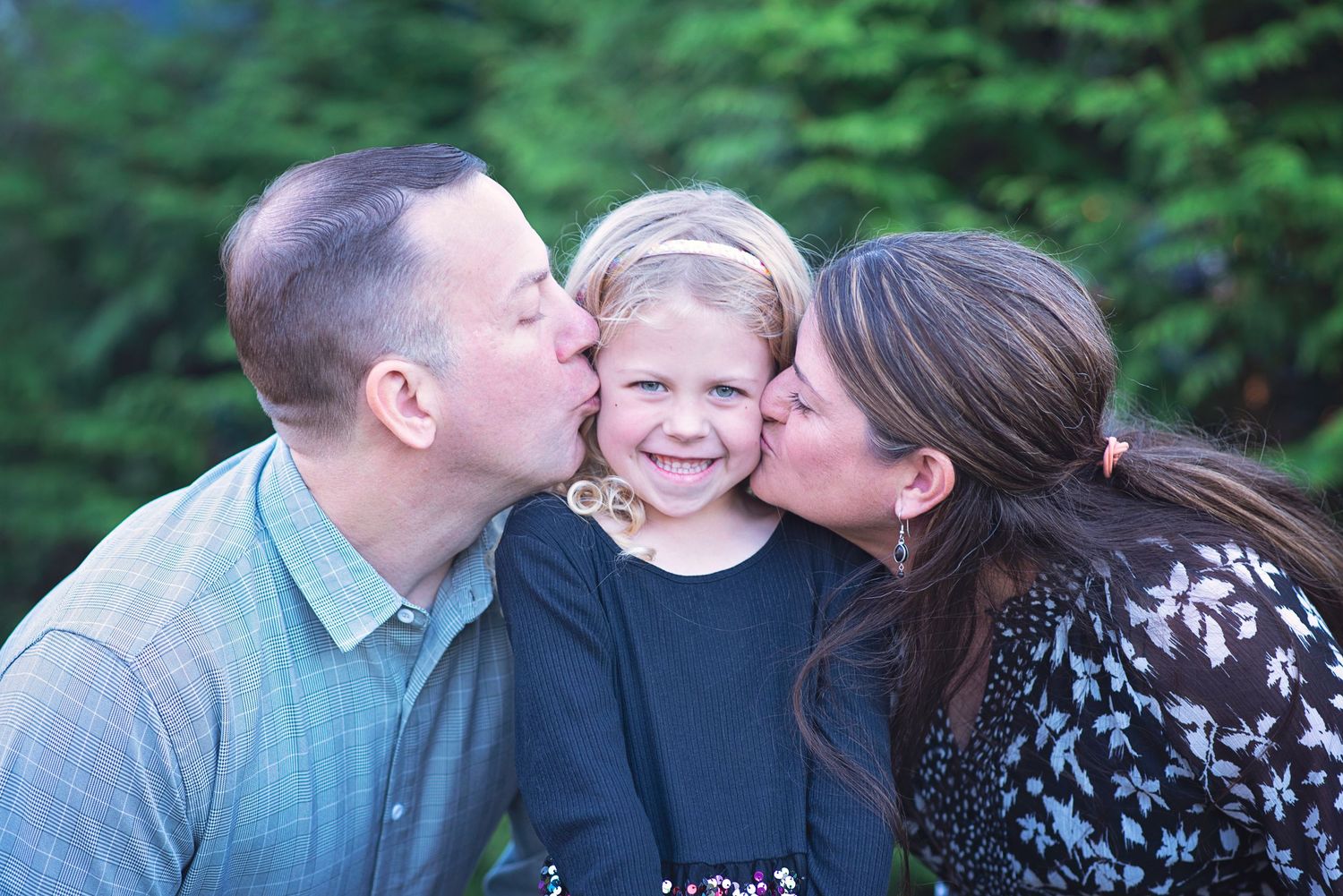 Parents give loving kisses to their smiling young child standing between them outdoors with green trees in background.