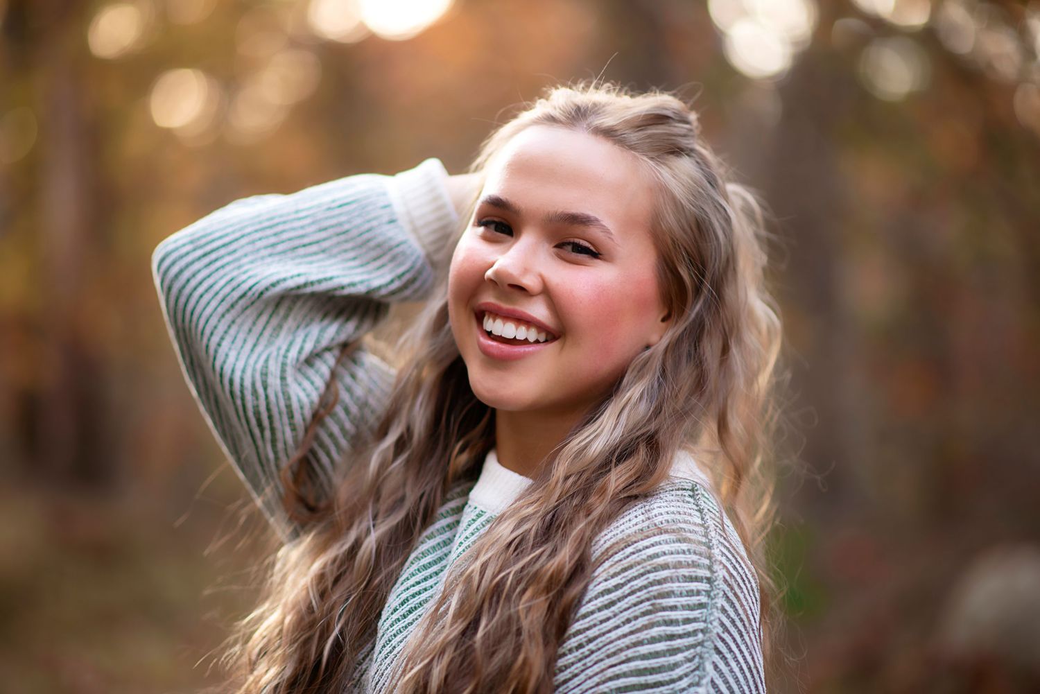 Young person with long wavy hair and gray sweater smiles outdoors with autumn bokeh background.