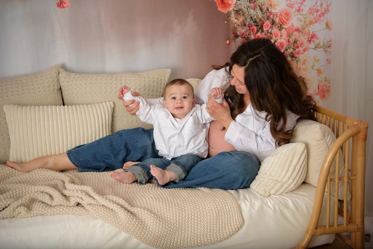 A mother and baby boy sit together on a cozy beige daybed with floral backdrop in a warm studio photo.