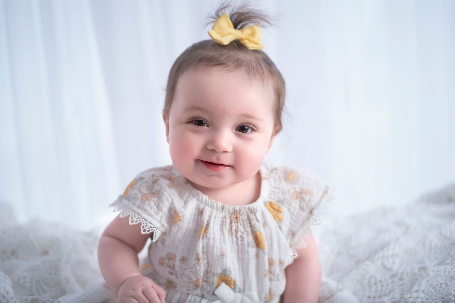 A smiling baby wearing a lace dress and yellow hair bow sits on white fabric.