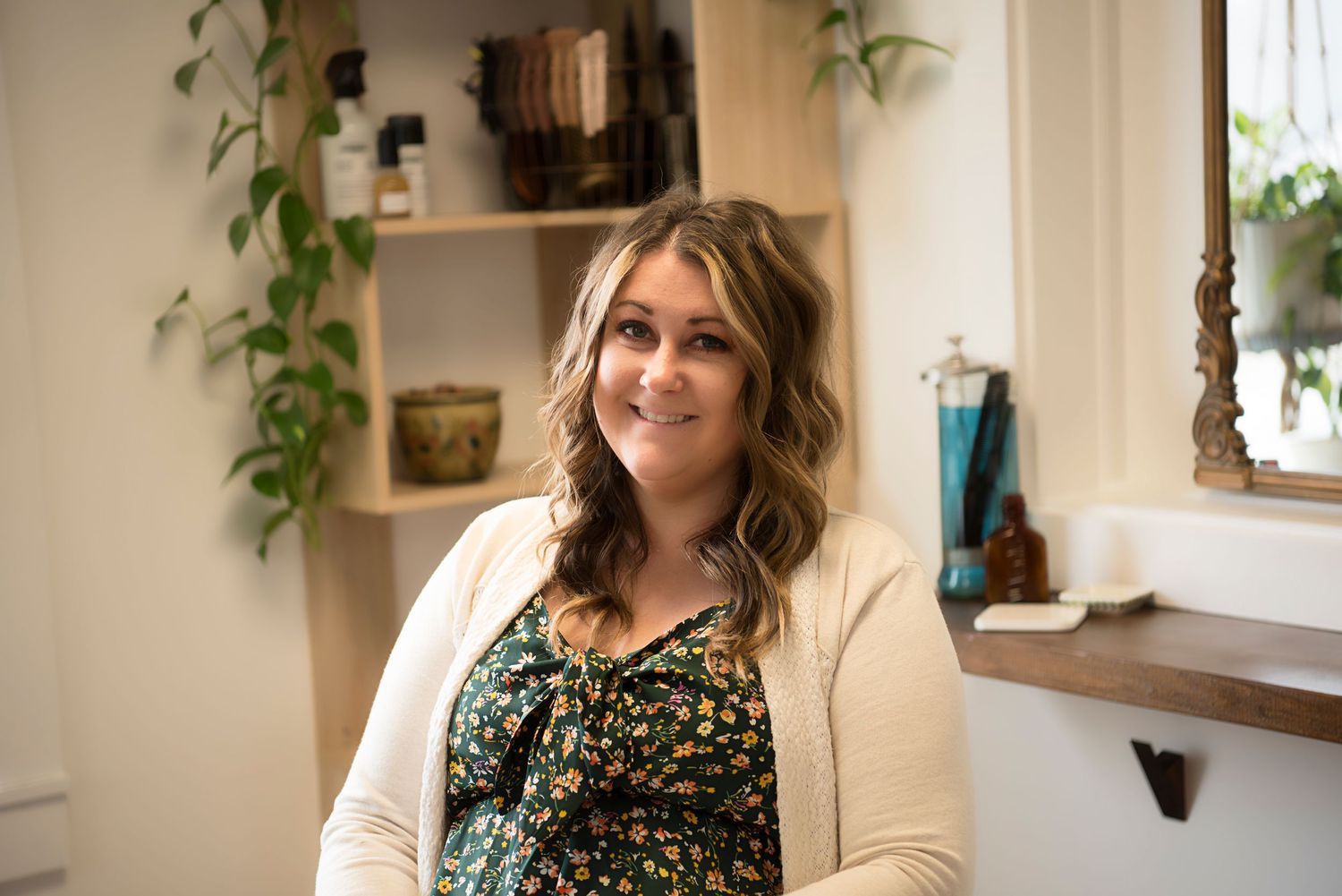 A person in a floral top and cream cardigan smiles while sitting in a cozy room with plants and shelves.