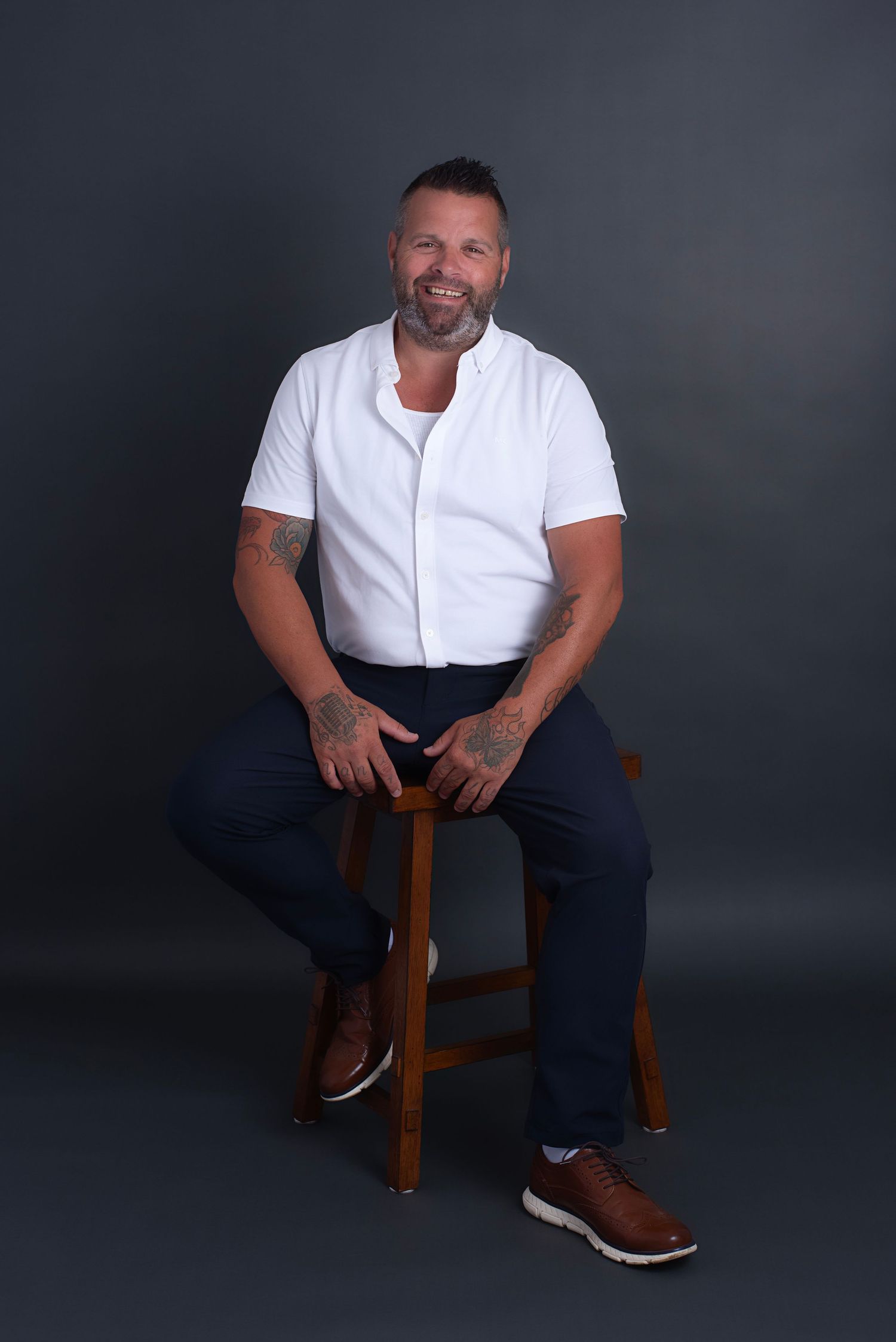 Person in white t-shirt and dark pants sits casually on wooden stool against dark background.