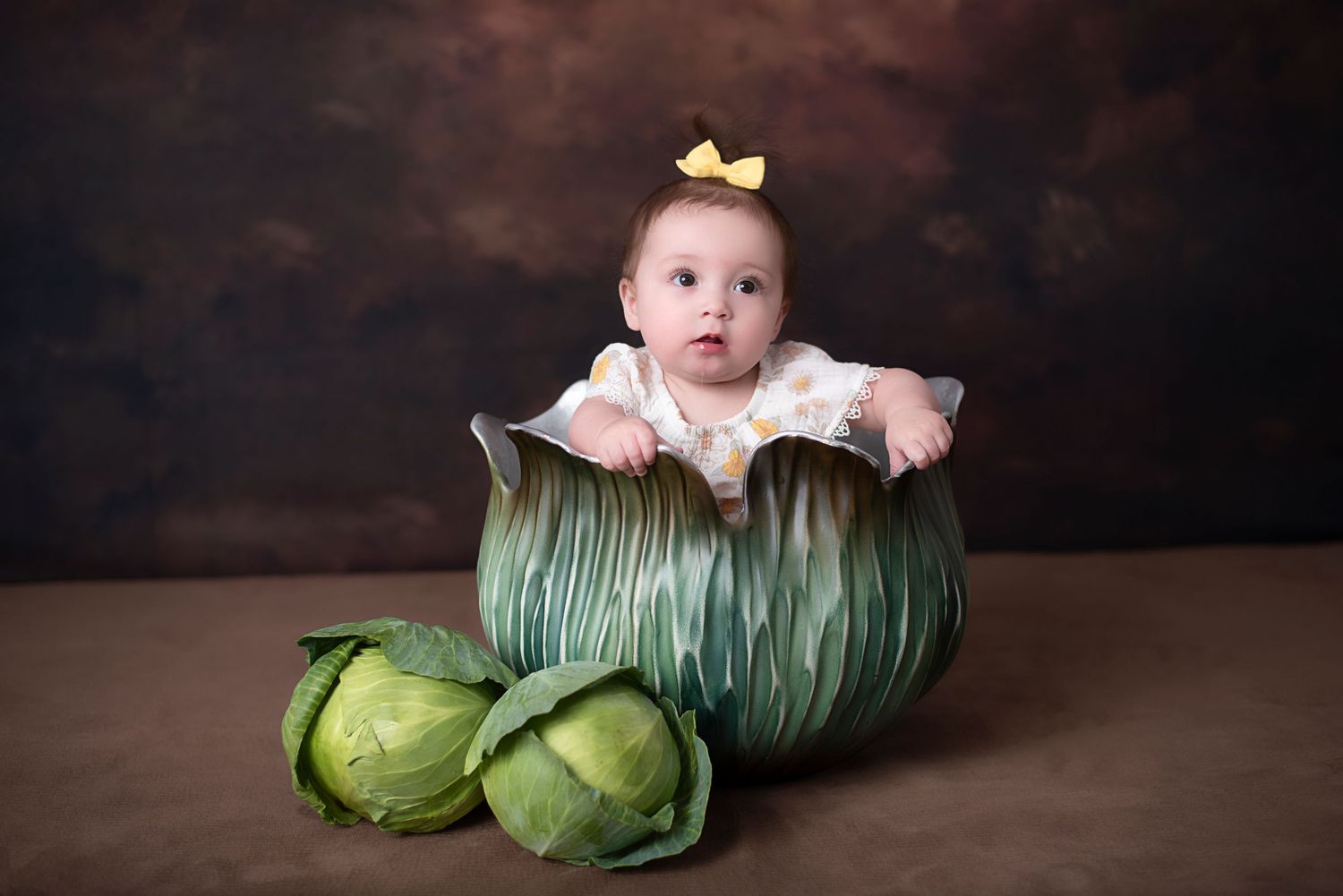 Baby wearing yellow bow sits inside giant decorative cabbage prop with real cabbages beside it in studio setting.