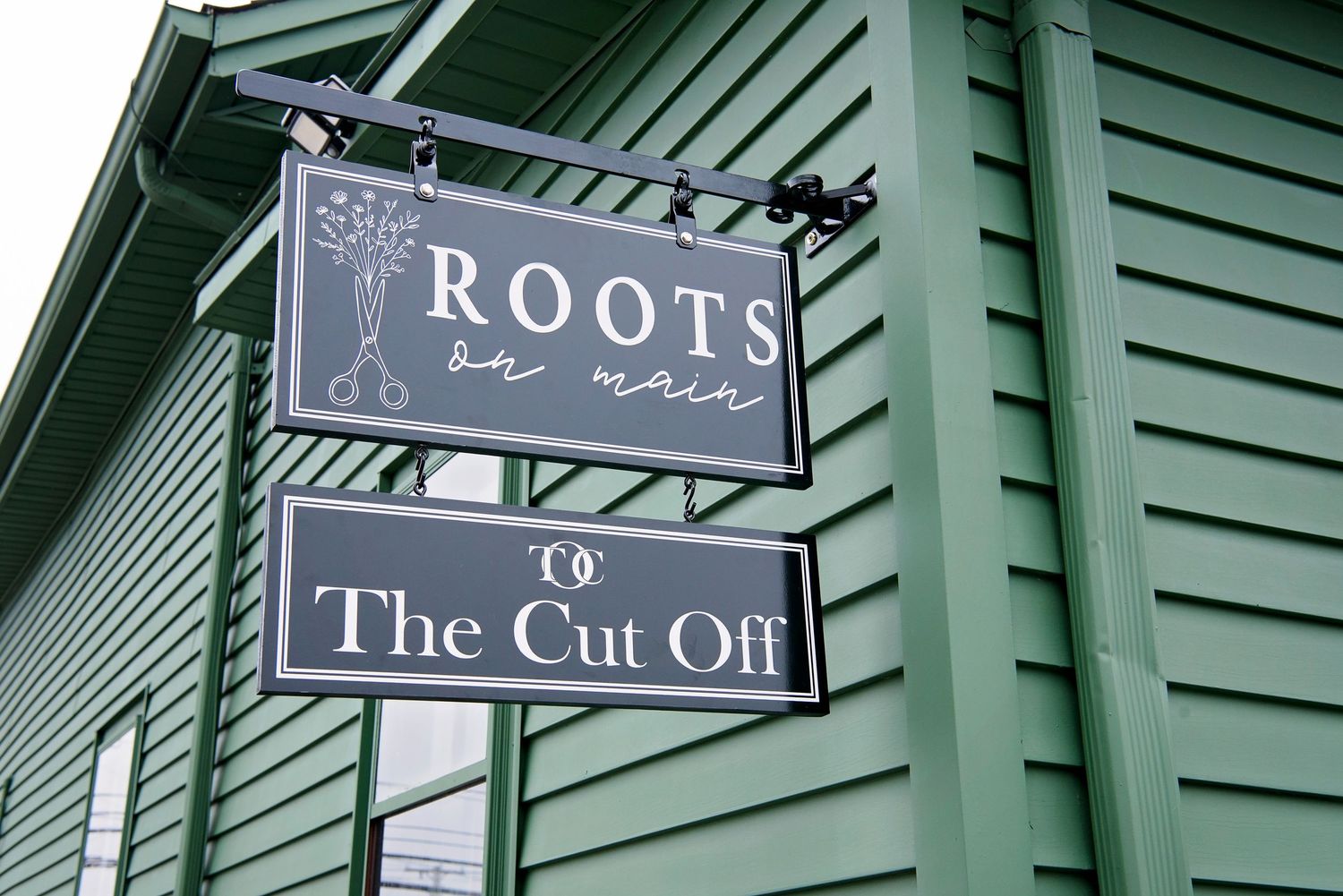 Two vintage style storefront signs advertising 'The Cut Off' and 'Roots' mounted on a green building exterior.
