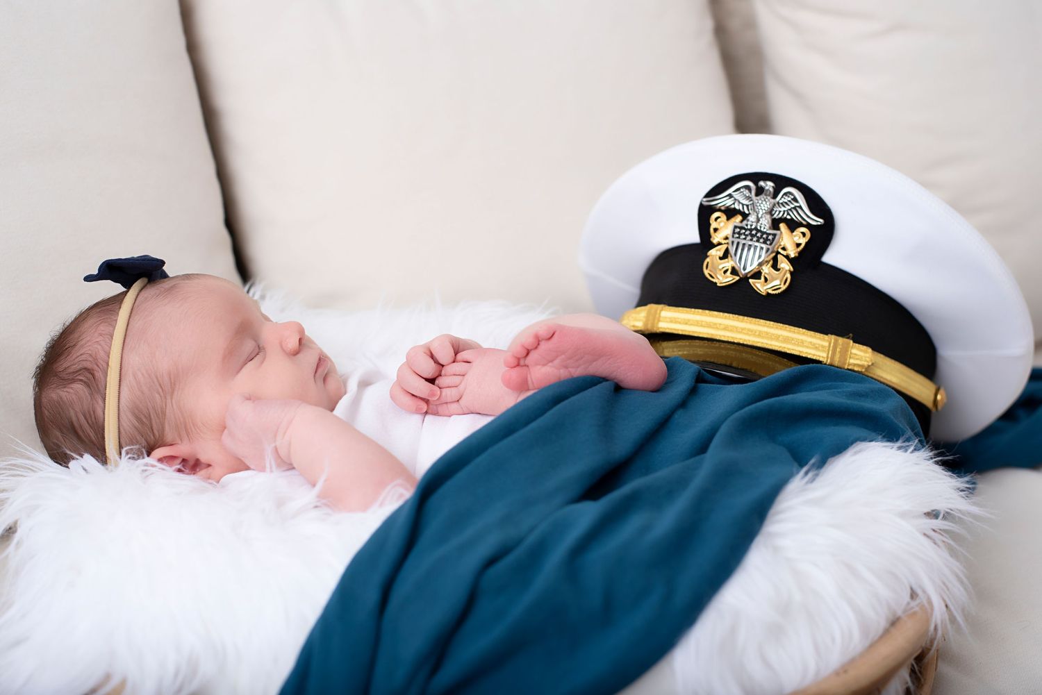 A baby sleeps peacefully while wearing a navy blue outfit and white military officer's hat with gold insignia.