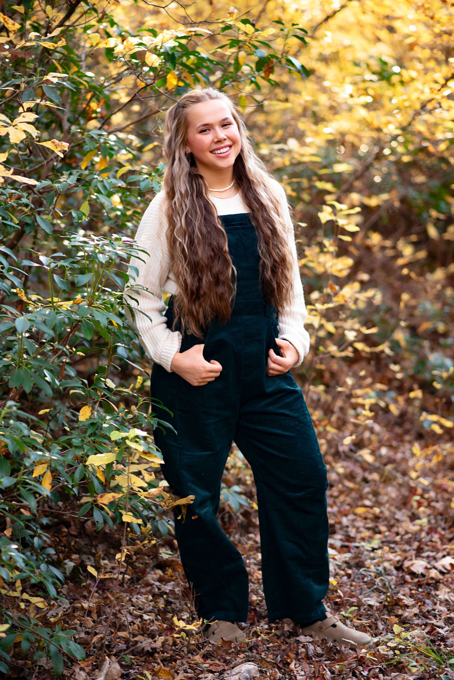 Someone poses outdoors wearing a black overall outfit with white sleeves against a backdrop of autumn foliage.