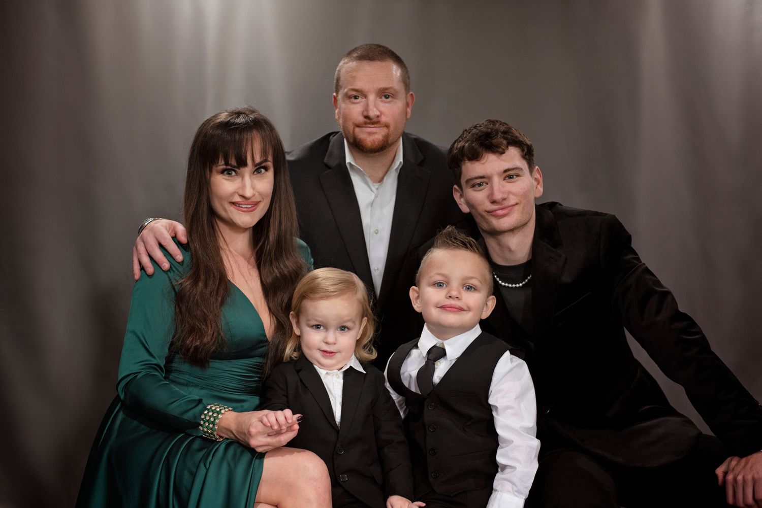 A formal family portrait with elegant attire featuring emerald green dress and suits against a dark gray studio backdrop.