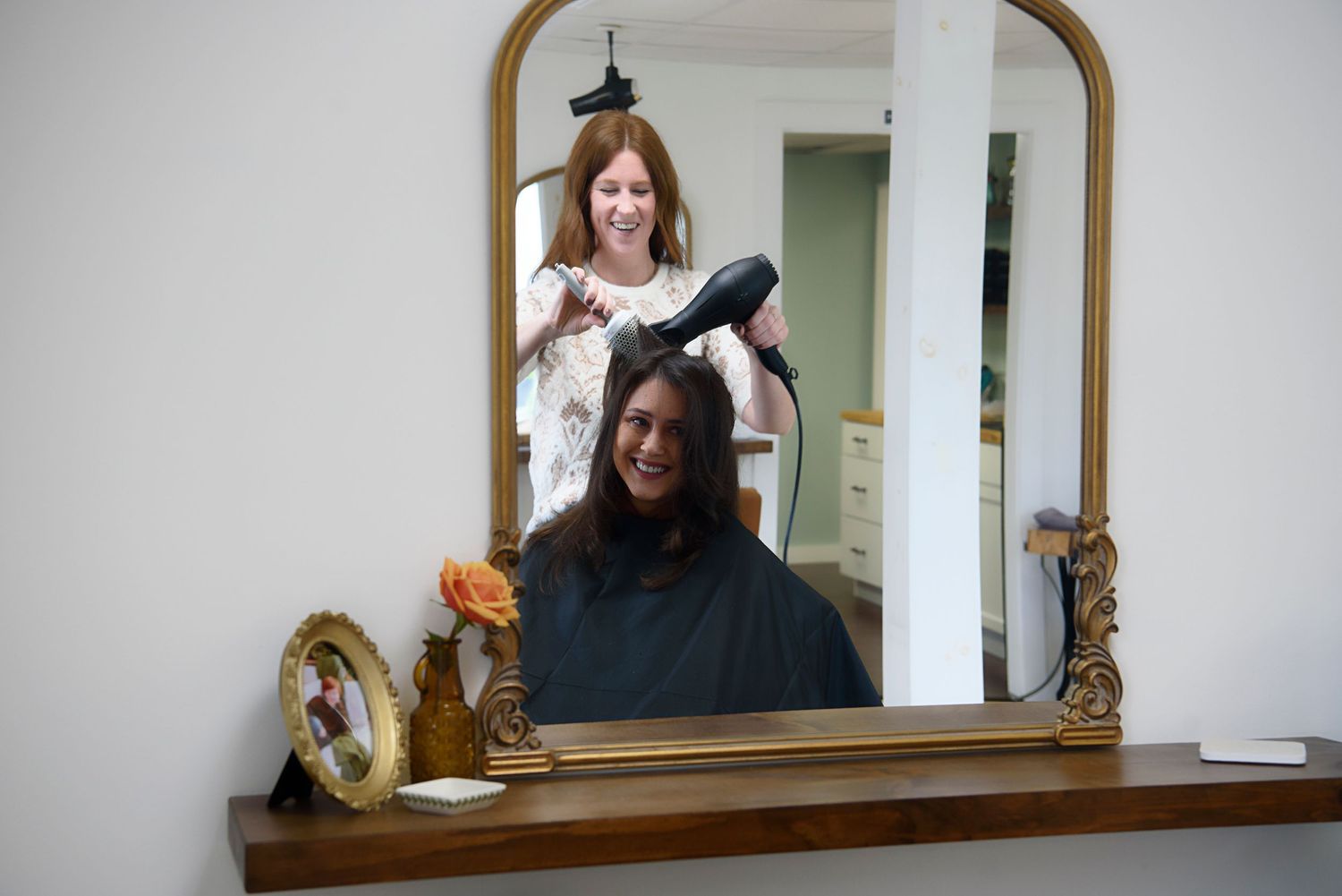 Hairstylist drying client's hair in salon with round mirror and decorative frame on vanity.