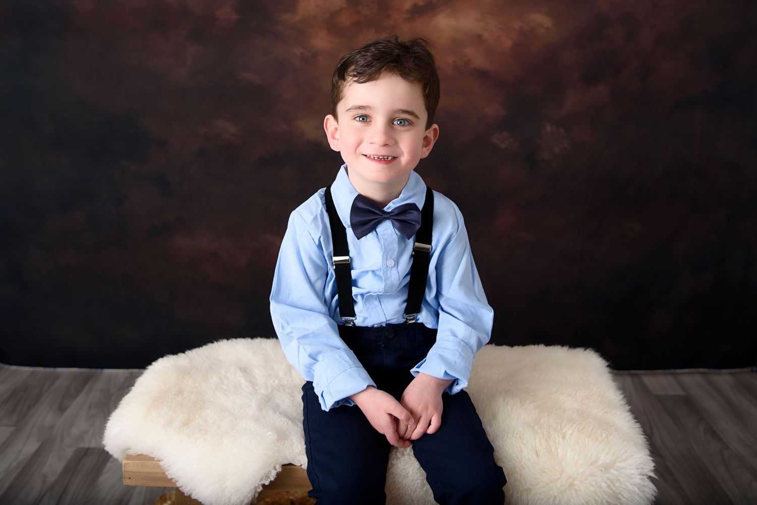 A boy in a blue dress shirt and bow tie sits cheerfully on white fur cushions against a dark background.