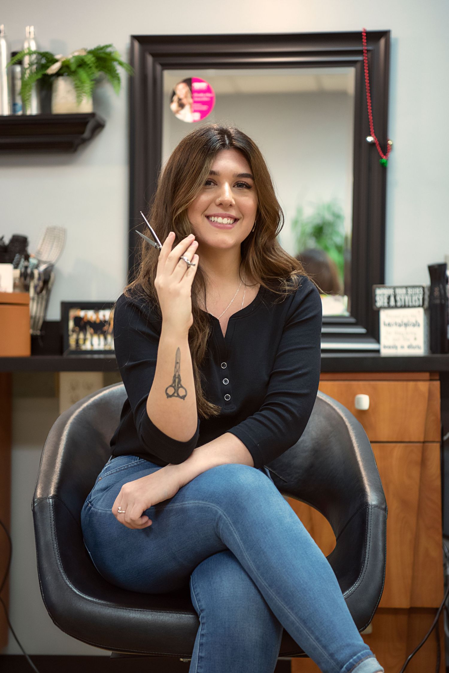 A hairstylist sits relaxed in a black salon chair wearing jeans and a dark top in a modern salon setting.