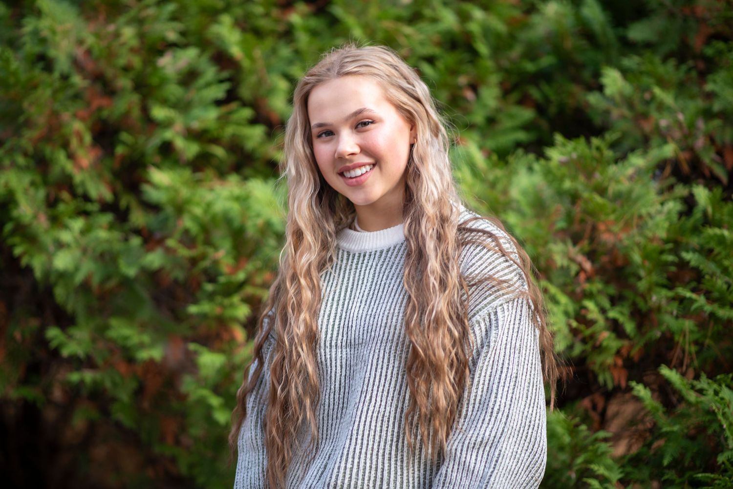 A smiling person with long wavy hair wearing a cozy grey sweater poses outdoors against green foliage. Rhode Island Photographer