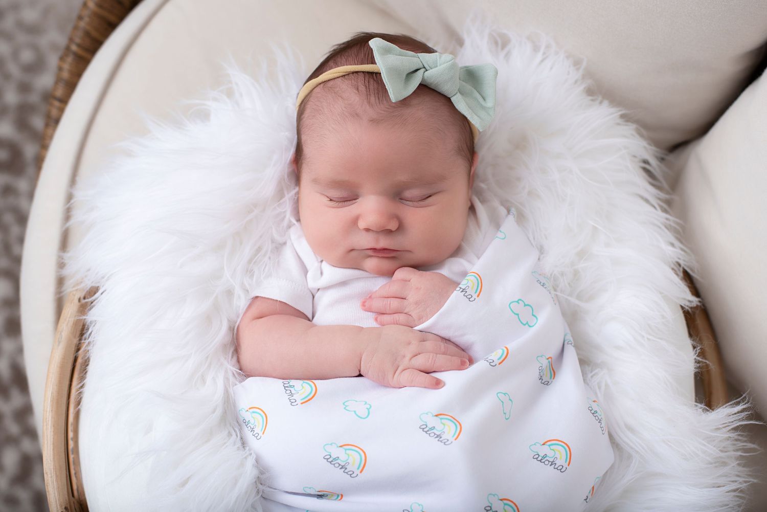 Sleeping baby with mint green bow resting on white furry blanket in basket with cloud printed swaddle.