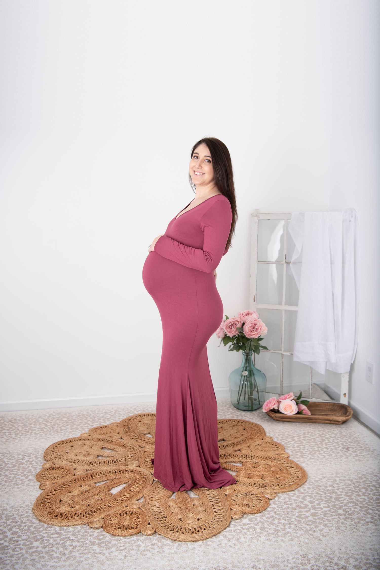 Expectant mother in flowing mauve gown poses in front of white wall with decorative rug and pink roses.