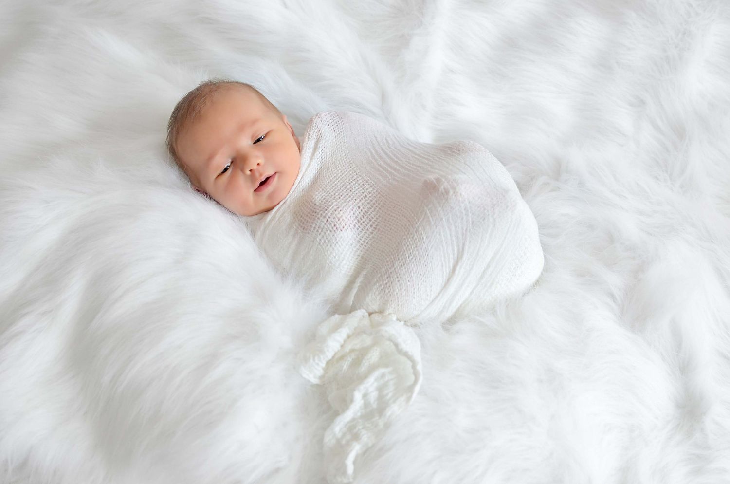 Newborn swaddled in soft white blankets rests peacefully against white fabric background.