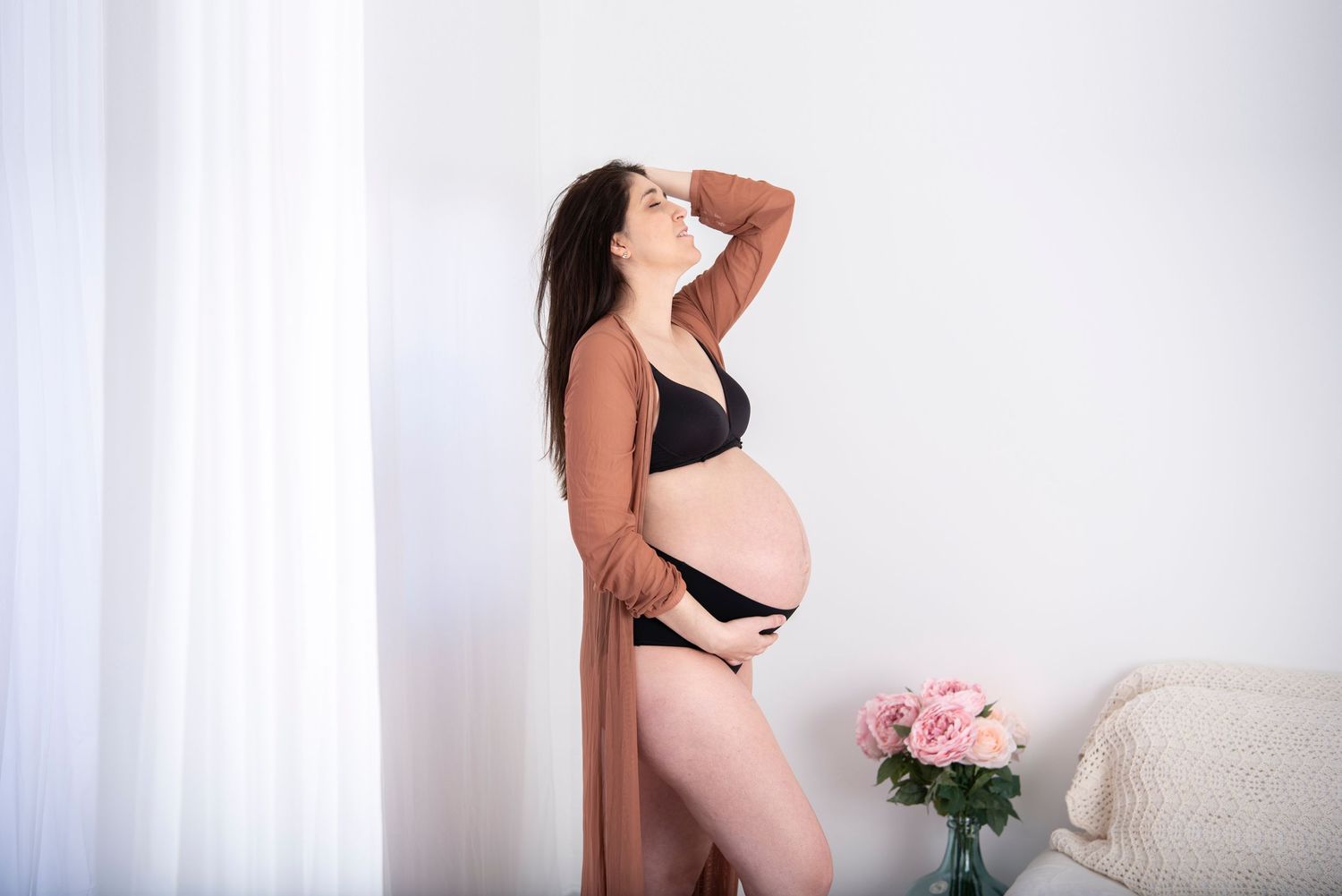 Person in black lingerie standing next to pink roses in a bright minimalist room.