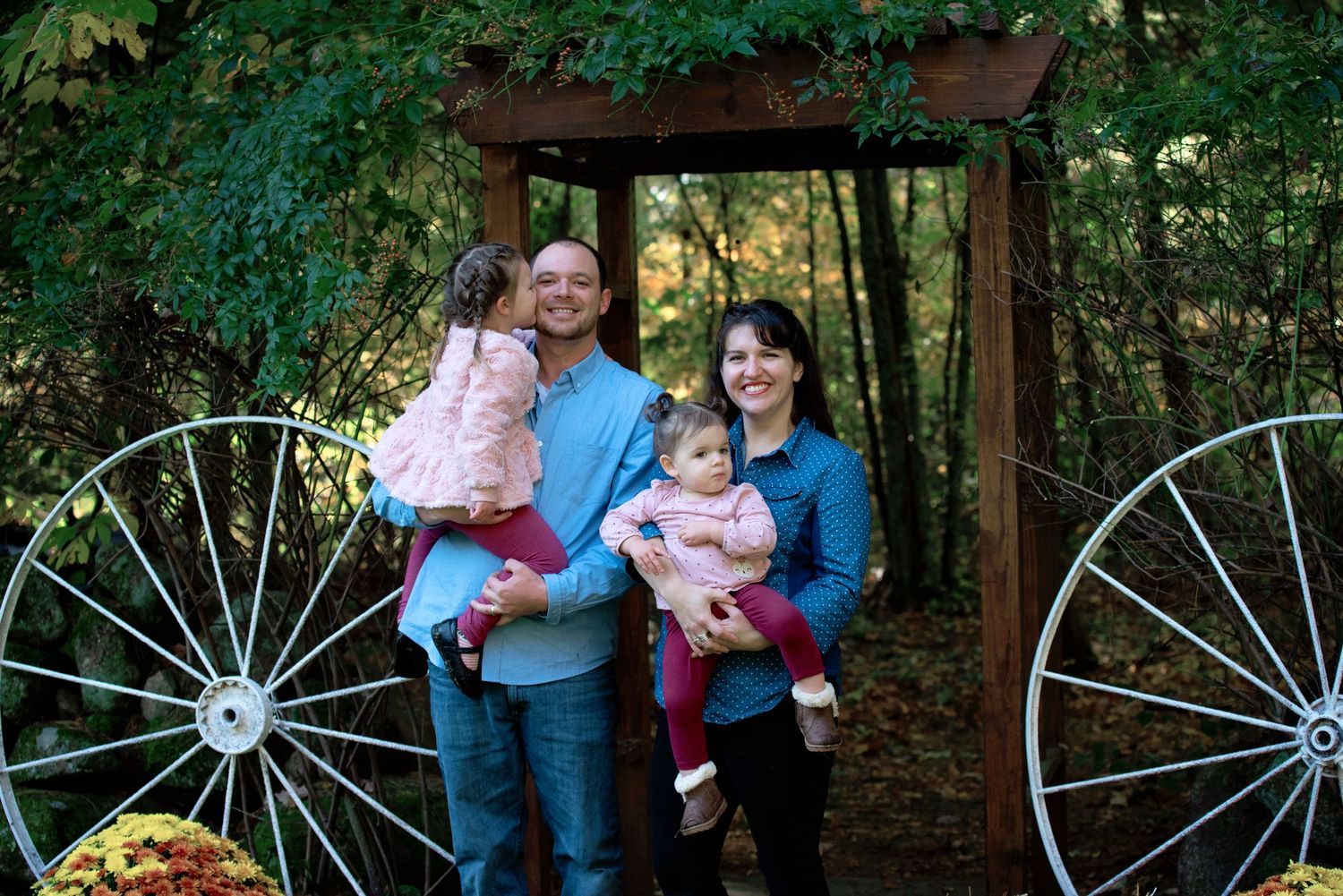 A family poses together under a wooden arch with wagon wheels and autumn decorations in a wooded setting.