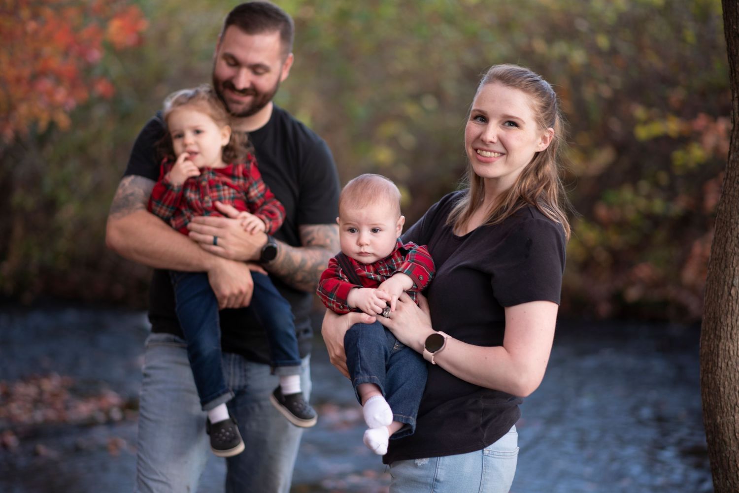 A family cuddles their twin babies outdoors in matching red outfits near a stream during autumn.