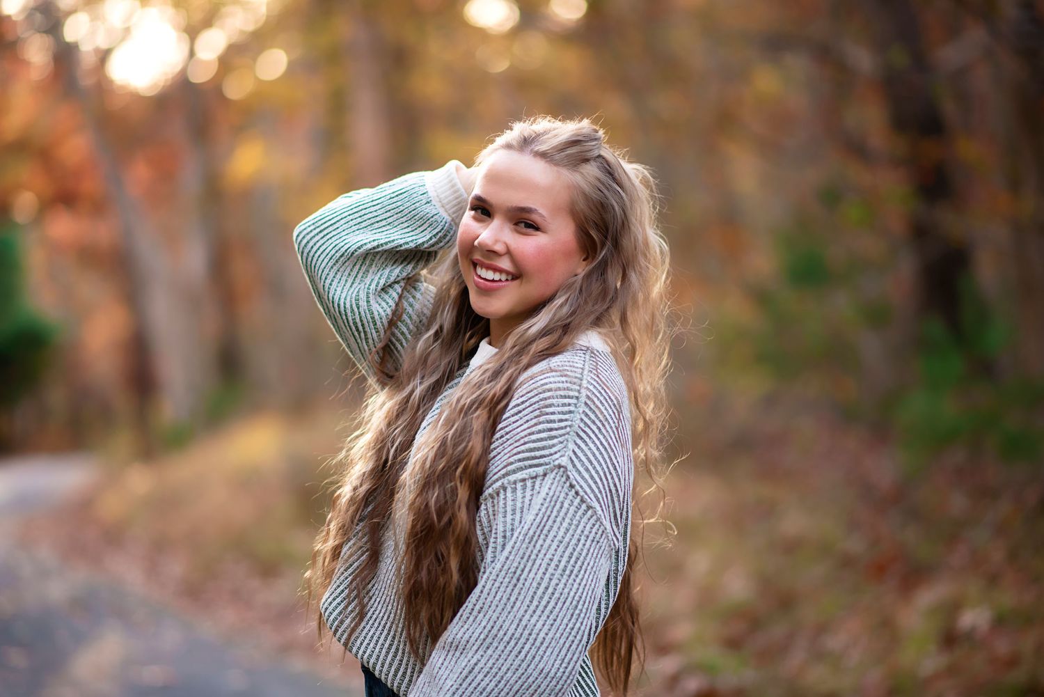 A smiling person with long wavy hair wearing a gray sweater poses outdoors during autumn with warm lighting. Rhode Island Photographer