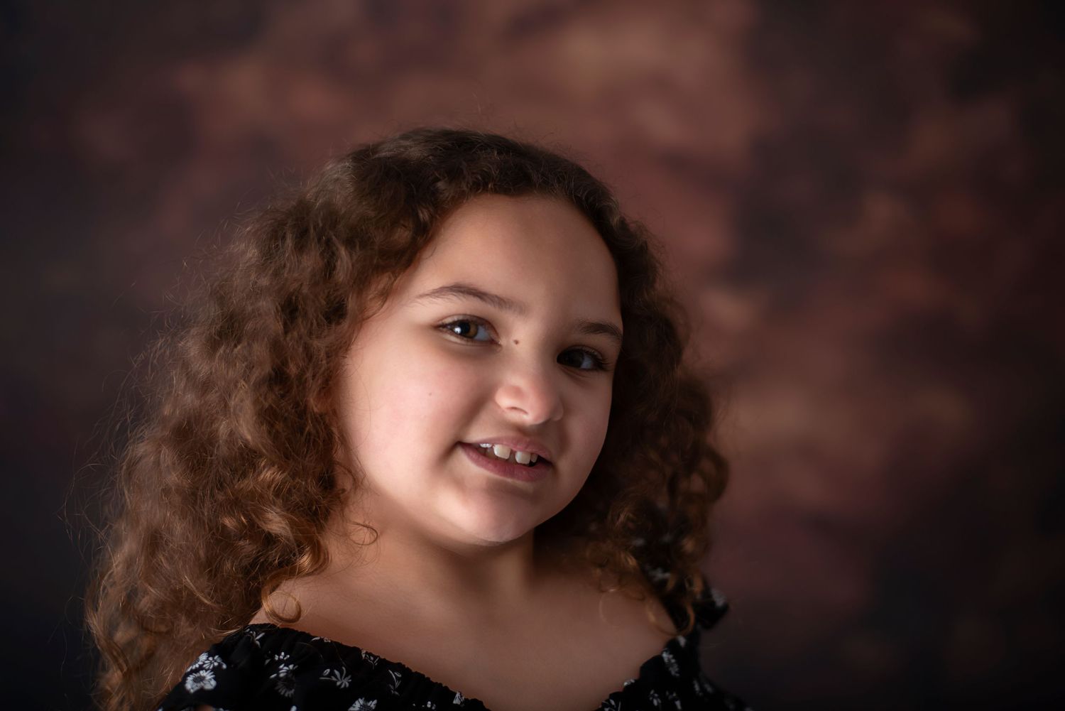 Young person with curly brown hair and a bright smile poses against a dark maroon studio backdrop.