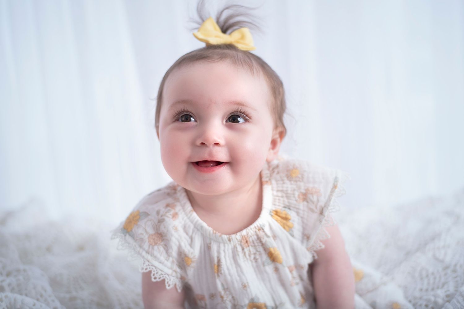Baby in white lace dress smiles brightly against soft white background with little blonde hair tuft on top.