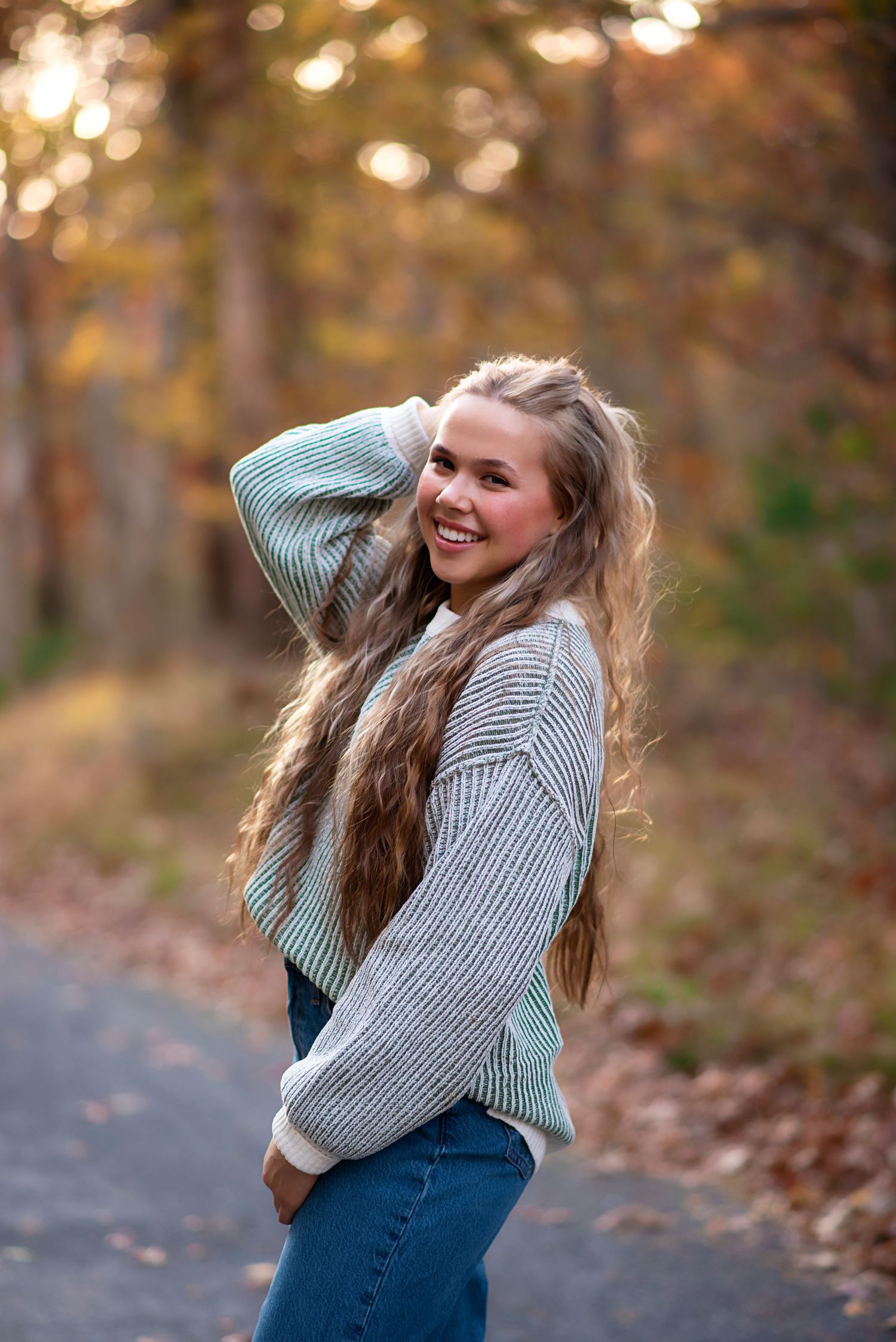 A person in a striped sweater and denim dress laughs while playing in autumn leaves during a photo session.