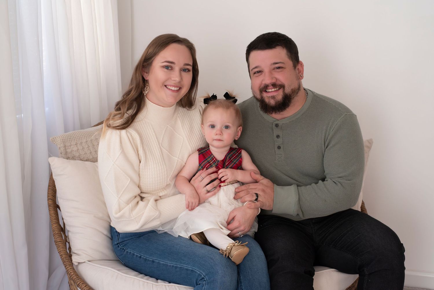 A family wearing neutral colors sits together on a couch for a series of casual indoor portrait photographs.