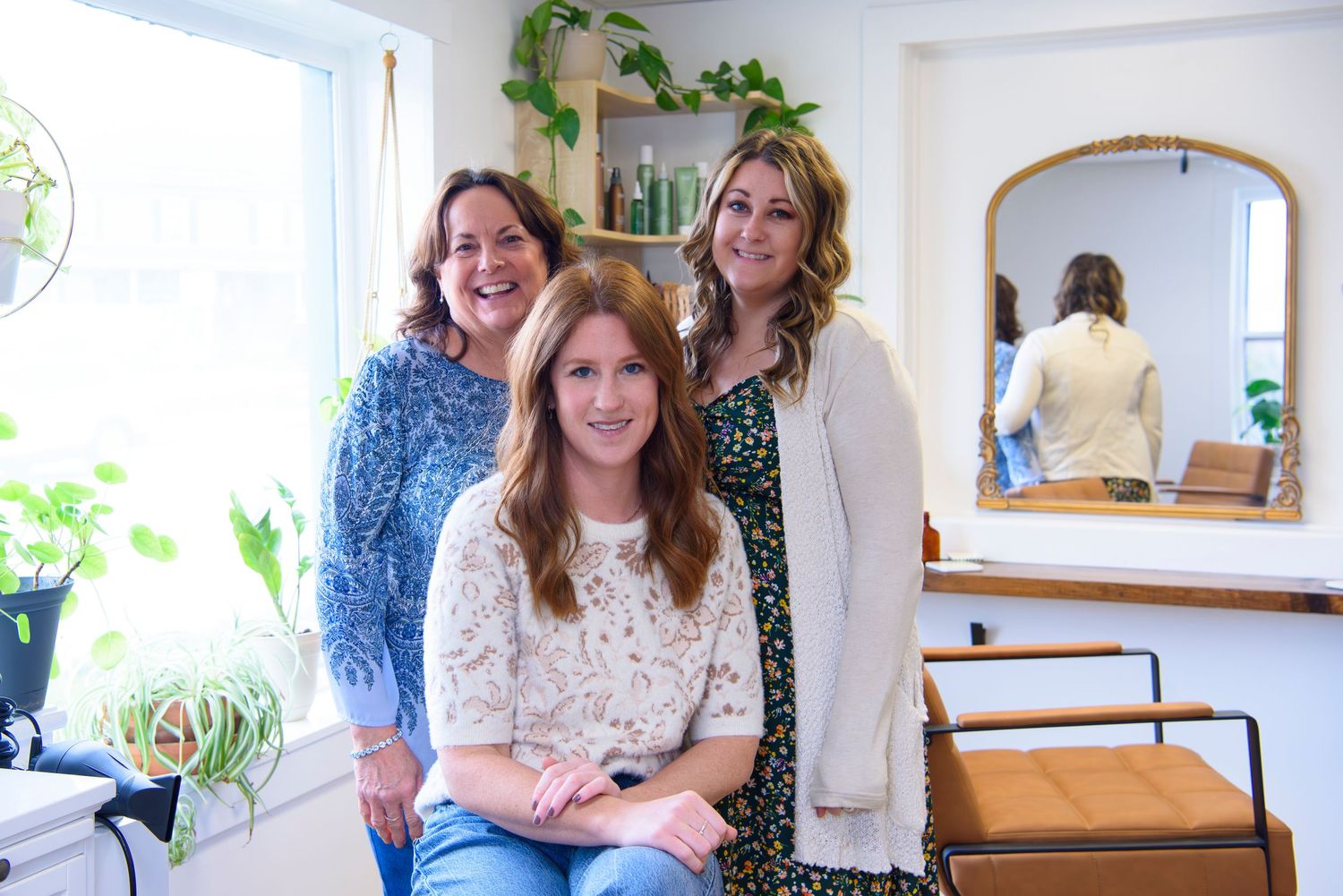 Three people sit and stand in a bright salon room with house plants and a wall mirror.