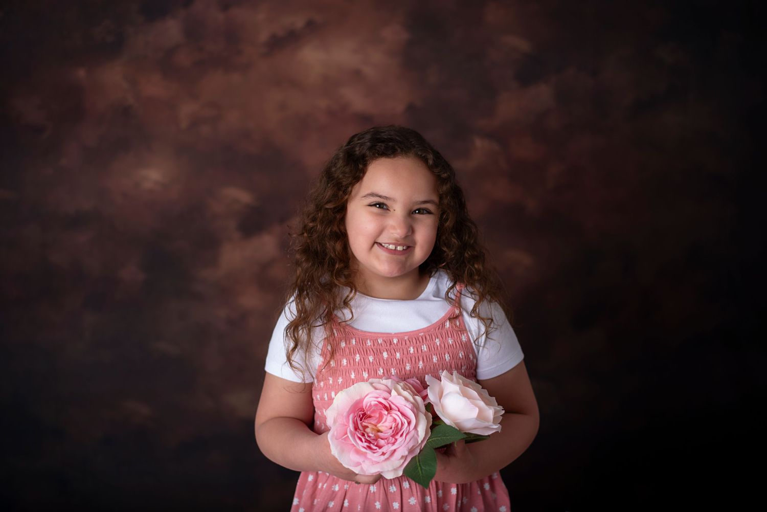 A curly-haired subject in a pink and white dress holds a single pink rose against a dark studio background.
