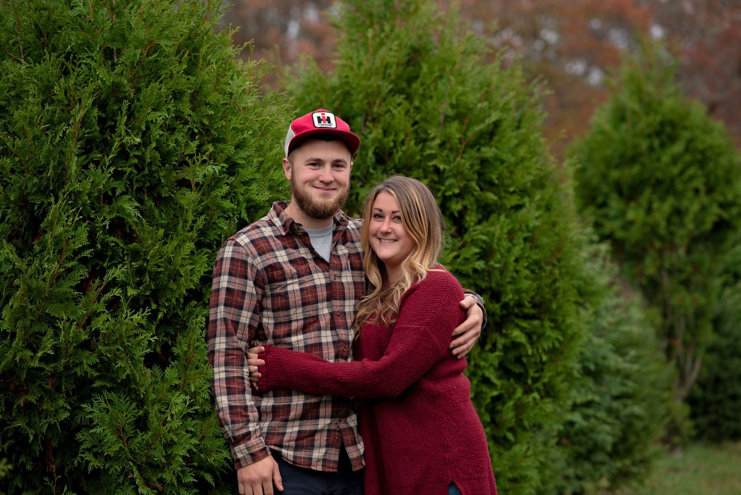 Two people in plaid and burgundy clothing pose together at a Christmas tree farm in fall or winter.