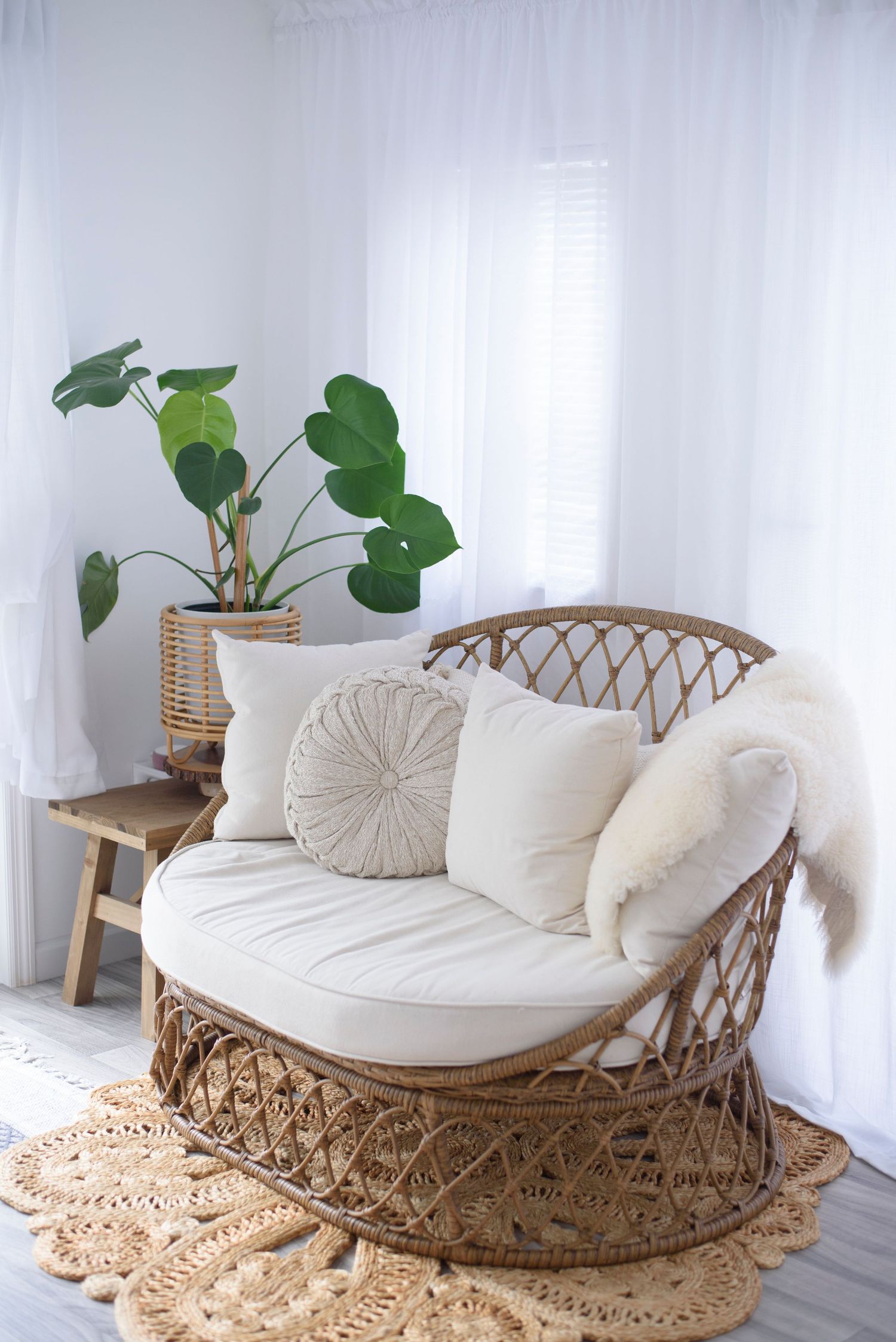 Cozy rattan loveseat with white cushions and pillows sits on a crochet rug beside a potted monstera plant in bright room.