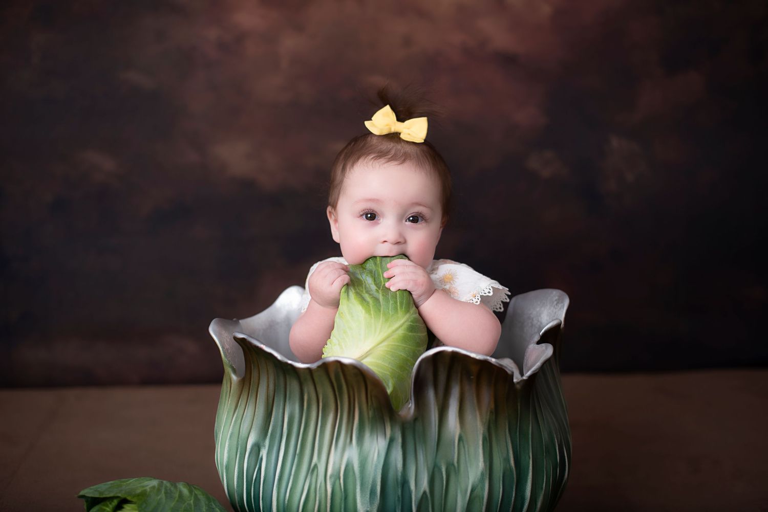 Baby wearing yellow bow sits inside decorative green cabbage prop during professional portrait session.