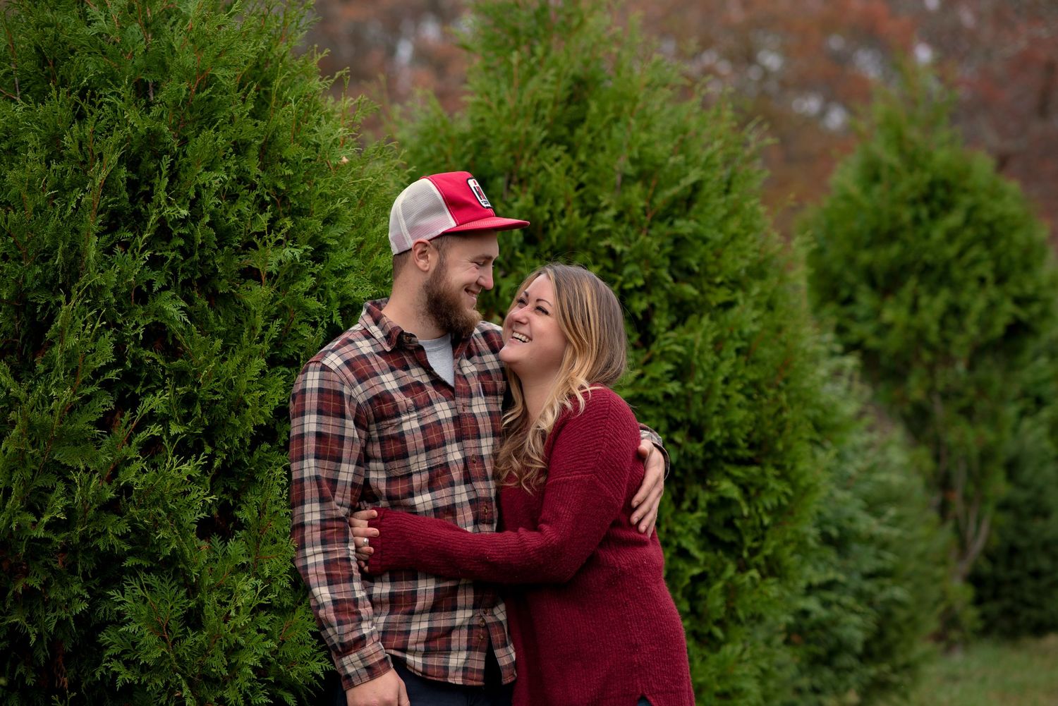 A couple in plaid and burgundy clothing share romantic moments in a garden surrounded by evergreen trees.