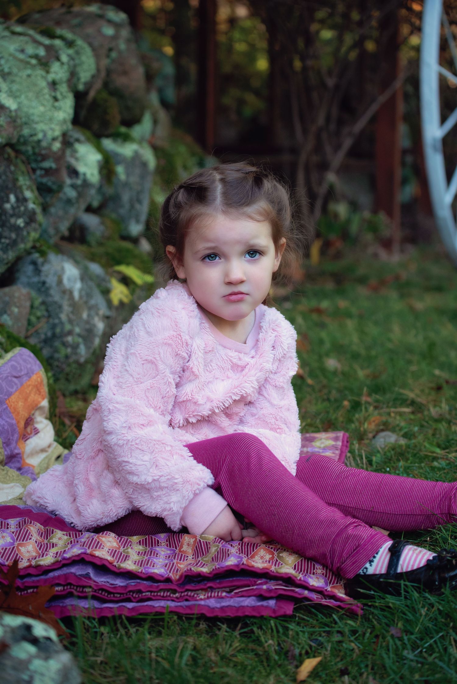 Child in pink fluffy sweater and purple tights sits on colorful blanket outdoors near rocks and foliage.