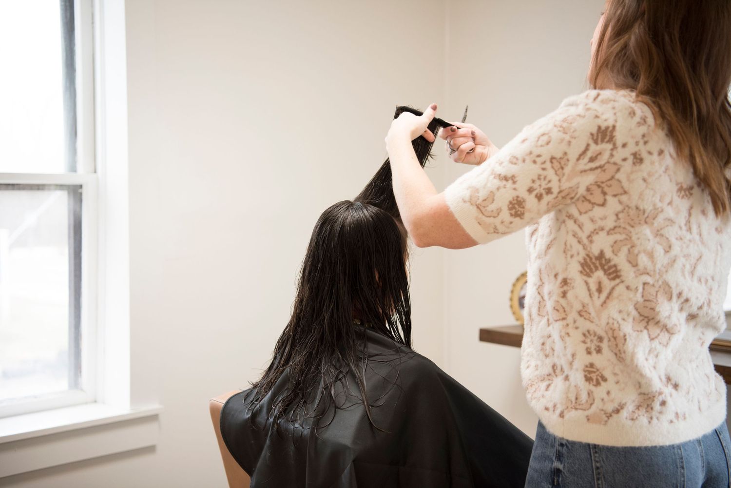 Professional hairstylist wearing white lace top cuts and styles dark hair in bright salon near window.