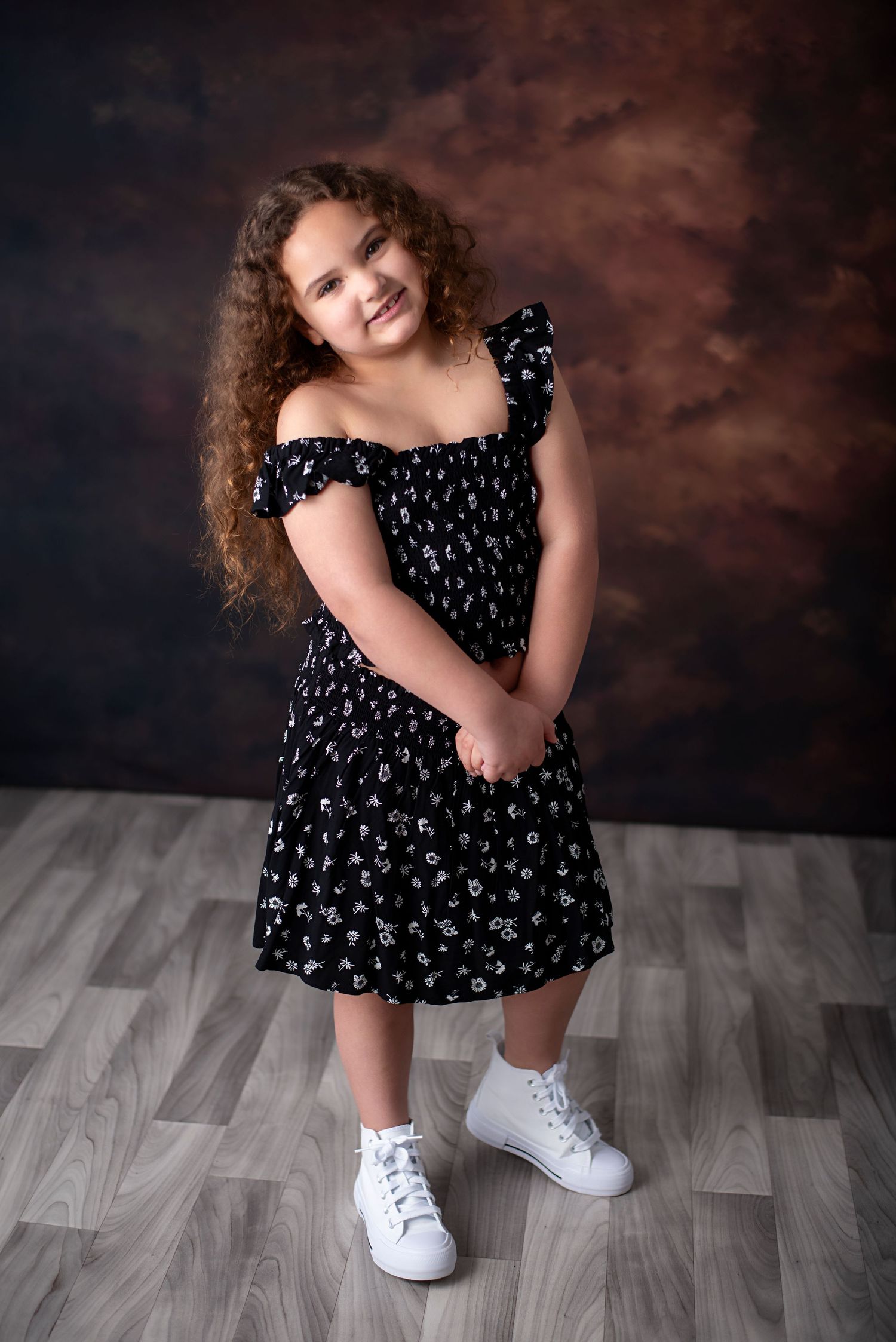 Portrait of someone in a black floral dress with white sneakers standing on gray wooden floor against dark background.